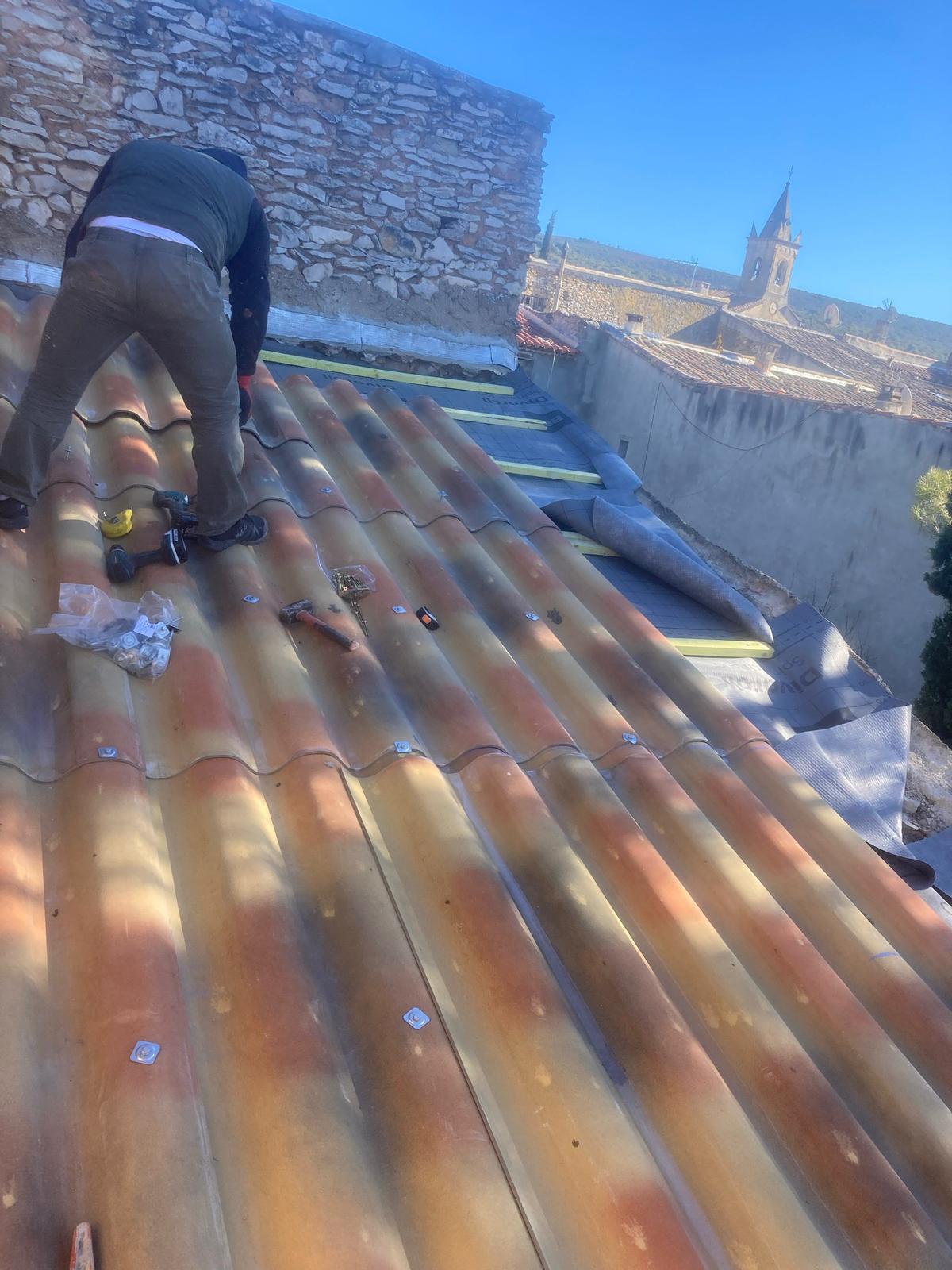 Dylan Scheid au travail sur un toit de village, devant un mur en pierres sèches et un clocher
